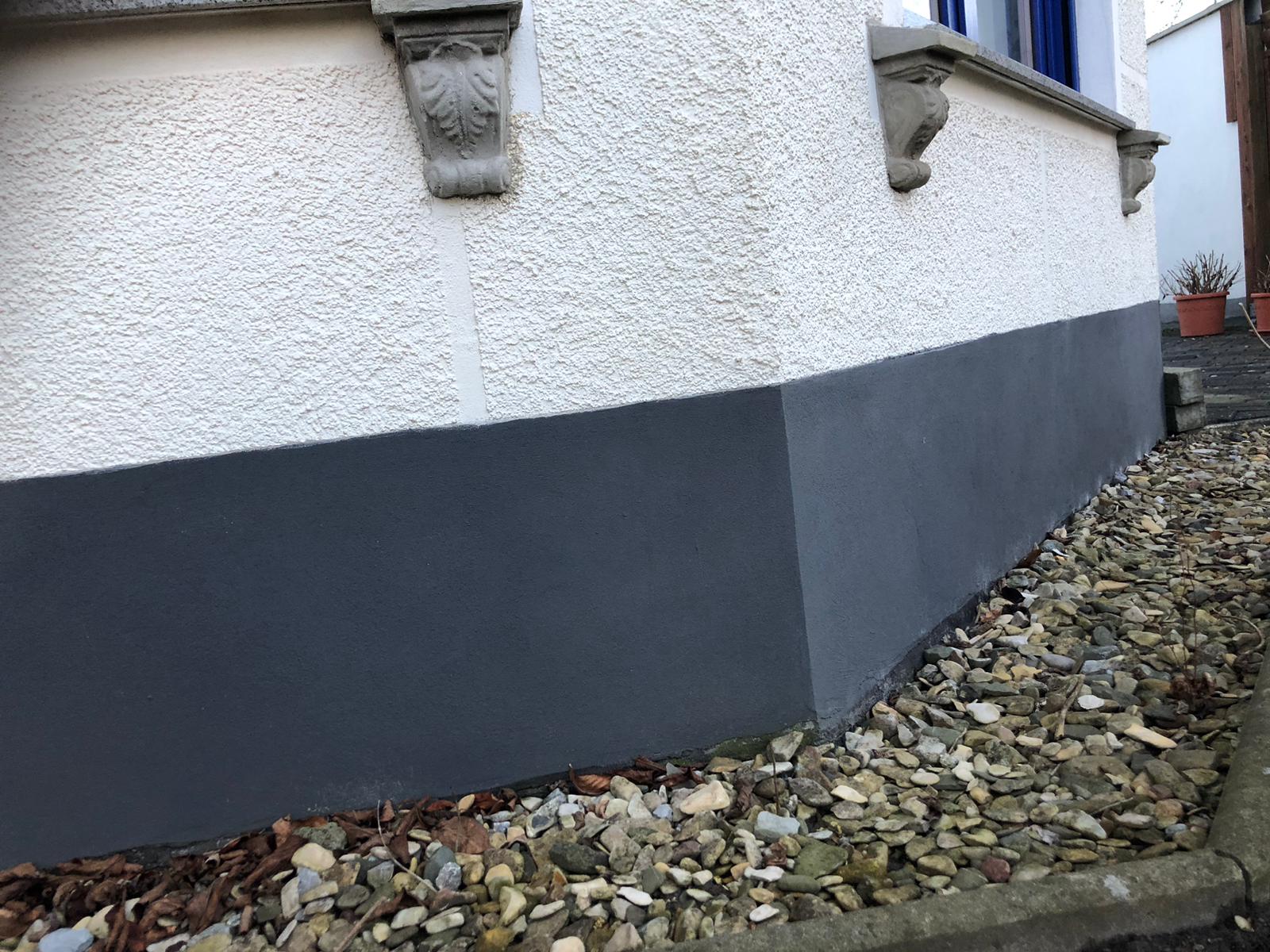 Außenfassade mit weißem Filzputz und dunkelgrau abgesetztem Sockelbereich, eingefasst von einer Kiesschicht entlang des Gebäudes